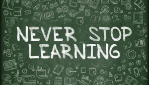 Green chalkboard with doodles of books, light bulbs, laptops, and learning symbols, featuring the bold white chalk text “NEVER STOP LEARNING” in the center.